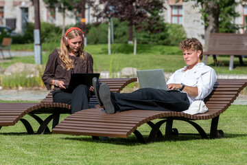 Obraz premium Man and woman sitting on reclining chairs and working with laptops in a modern urban tech hub outdoor area in the summer