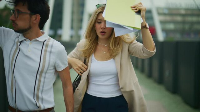 Business colleagues struggle with strong wind while walking outdoors, trying to protect their documents. A man and woman in business casual attire are surprised by bad weather and battle the wind.