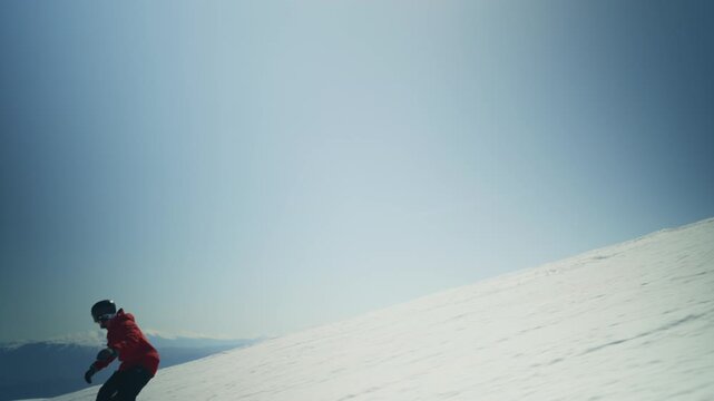 A person in a red jacket and helmet is snowboarding gracefully down a pristine white mountain. The snowboarder carves through the snow under a clear, bright blue sky.
