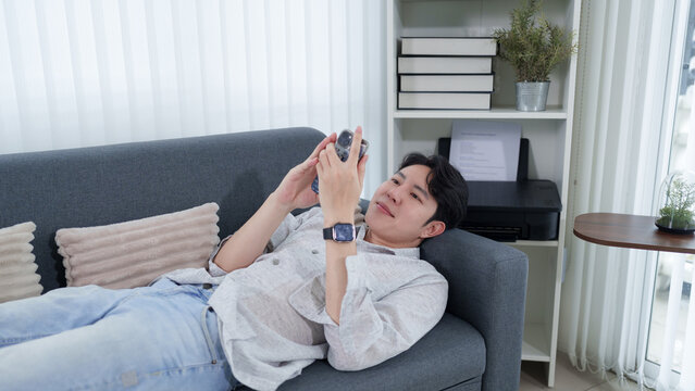 Young Asian man relaxing on sofa browsing smartphone at modern home living room with copy space