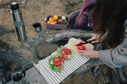 Woman slicing fresh tomato while preparing vegetable sandwiches for an outdoor lunch in the forest with a thermos, food container and backpack nearby