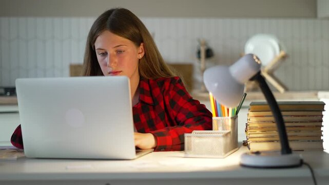 Focused teenage girl studying online using silver laptop. Young girl sits at bright desk doing school homework. Diligent student types on laptop keyboard. Hard homework requires deep studying.