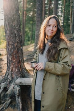 Woman holding a metal cup during a relaxing break in a sunlit forest, enjoying a warm drink and a peaceful moment of cozy outdoor lifestyle in nature