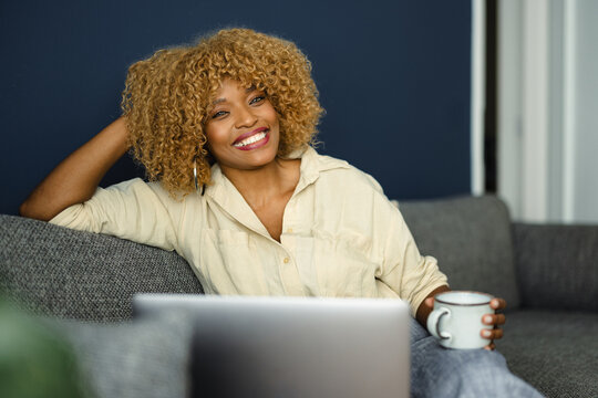 Woman Smiles While Sitting on Couch With Laptop and Coffee Cup