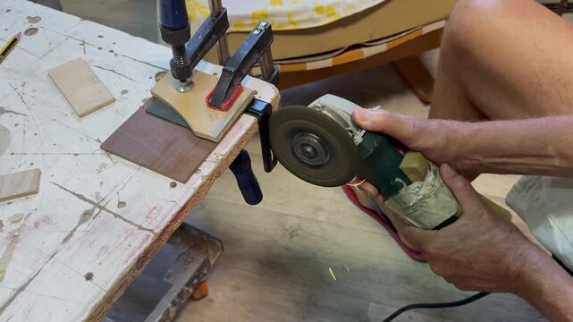 A side view of a craftsman using an electric power grinder to cut a steel bracket securely clamped to a workbench. Bright golden sparks illuminate the industrial workshop studio setting.