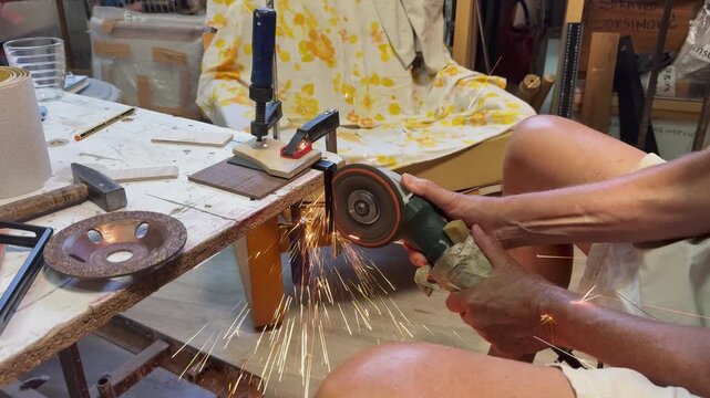 A side view of a craftsman using an electric power grinder to cut a steel bracket securely clamped to a workbench. Bright golden sparks illuminate the industrial workshop studio setting.