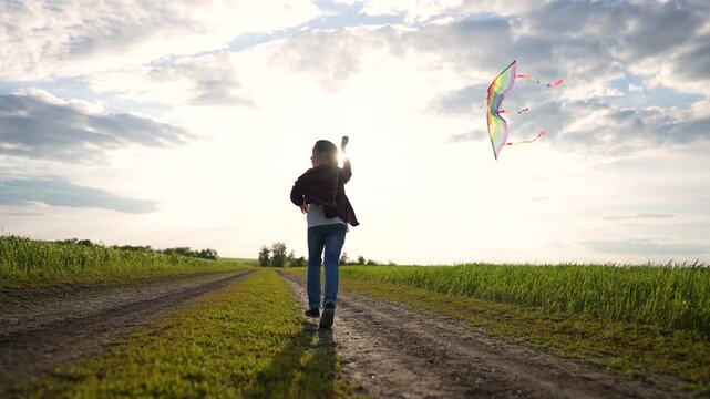 Child runs with rainbow kite along rural road at sunset. Small dog follows kid across green field. Happy girl plays outdoors in countryside. Carefree childhood freedom in nature at dusk.