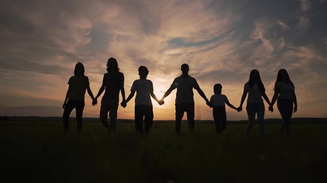 Large family walking forward holding hand. Dark silhouette stands against bright sunset sky. Group enjoys calm nature togetherness. Happy family walking on field. Group silhouette at warm sunset.