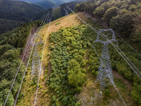 Aerial view of high-voltage power transmission towers in the Korytnicka dolina valley, surrounded by dense green forests and rolling hills in Liptovska Osada, Zilinsky kraj, Slovakia.