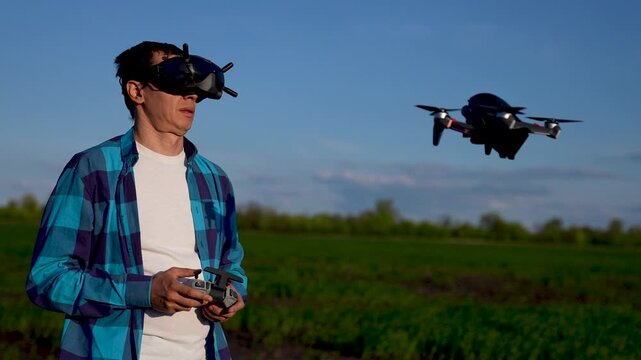 Man flying drone with FPV goggles in green field. Pilot controls drone with remote controller outdoors. FPV drone hovers near pilot at sunset. Man operates flying drone using goggles and controller.