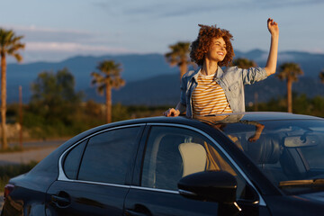 Fototapeta premium Joyful young woman leans out of a car window, raises her fist, as the sunset lights palm trees and distant mountains on a warm evening road trip.