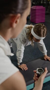 Vertical over the shoulder shot of young adult Caucasian woman trainer timing Caucasian child holding plank on step platform. Footage represents youth fitness programs and personal training services