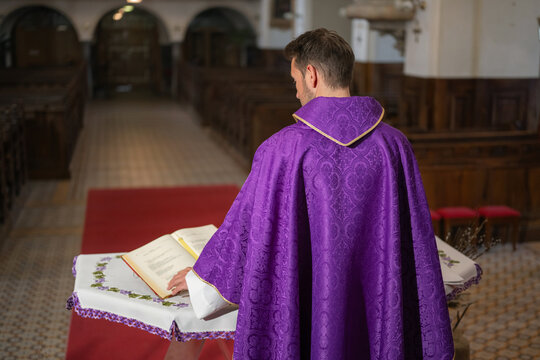 A priest stands in a church reading from a bible. The church has wooden benches and a red carpet.