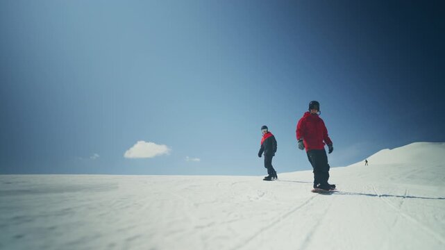 Two snowboarders glide down a pristine snowy mountain slope under a clear blue sky. They enjoy a sunny day while snowboarding at the ski resort.