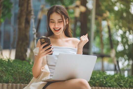Excited woman celebrating good news on smartphone with laptop outdoors, success and achievement concept