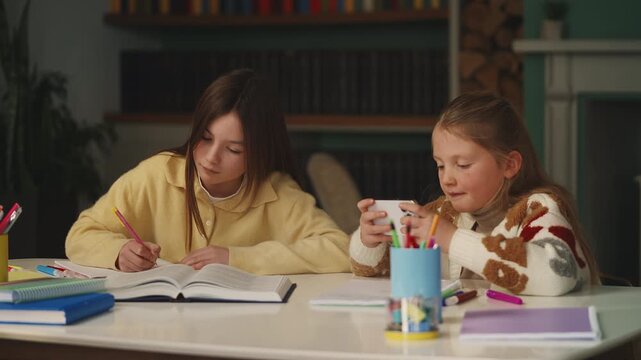 A girl plays mobile game while her diligent sister does homework nearby