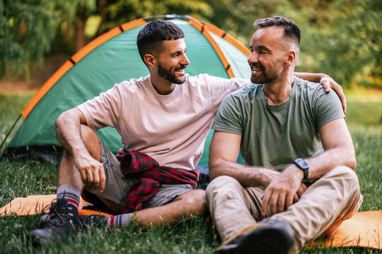 Gay couple sitting close together and smiling beside their camping tent
