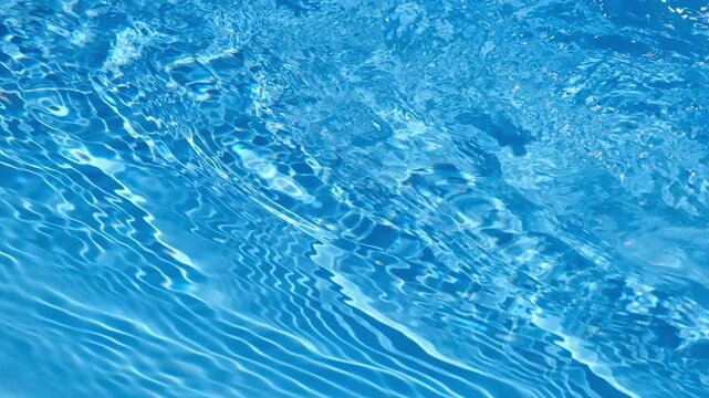 Clear blue swimming pool water surface with moving ripples, waves, reflections, and shimmering sunlight patterns.