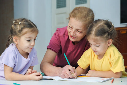 Smiling Grandmother Guides Two Little Girls While They Draw Together At Table With Colored Pencils And Open Notebook In Bright Home Interior, Showing Patient Family Support