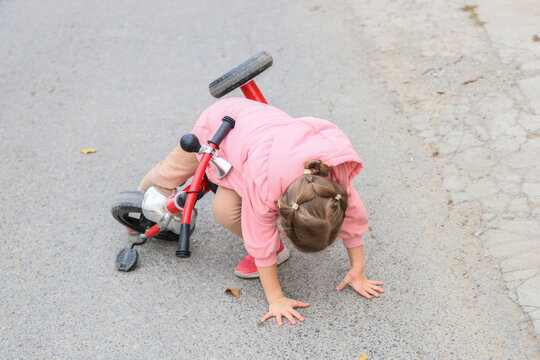Young Child In Pink Jacket Falls Beside Red Balance Bike On Paved Road, Hands On Ground, Showing Early Cycling Practice, Balance Struggle, Outdoor Learning Moment, And Childhood Determination