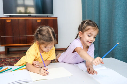 Two Young Girls Focus On Coloring And Sketching At Indoor Table, Using Paper And Pencils During Playful Art Activity In Bright Living Room With Modern Furniture