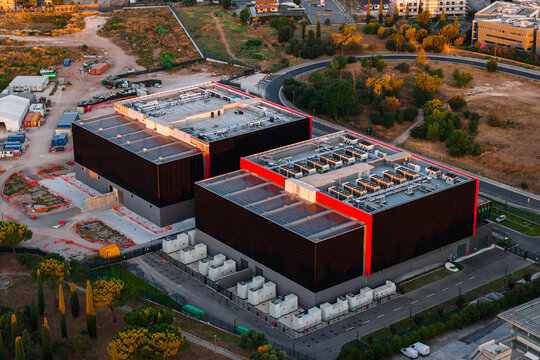 Aerial view of a modern datacenter complex with two large dark-walled buildings featuring rooftop HVAC units, surrounded by roads and greenery in Rome, Lazio, Italy.
