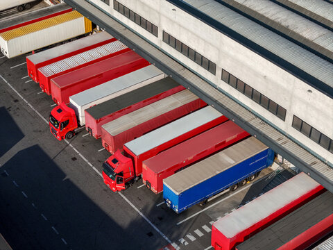 Aerial view of a logistics warehouse with multiple freight trucks parked at loading docks in an industrial yard in Settimo Torinese, Piedmont, Italy.