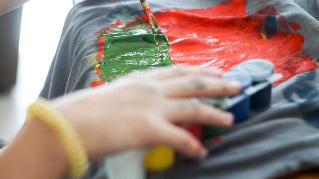 Child hands painting with acrylic colors on a grey t-shirt at home workspace