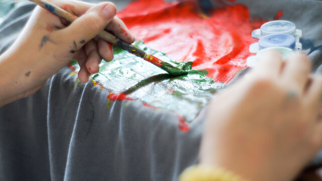 Child hands painting with acrylic colors on a grey t-shirt at home workspace
