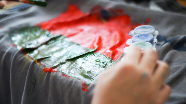 Child hands painting with acrylic colors on a grey t-shirt at home workspace