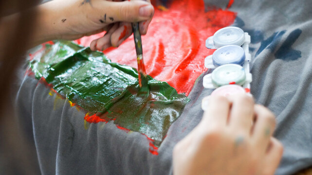 Child hands painting with acrylic colors on a grey t-shirt at home workspace