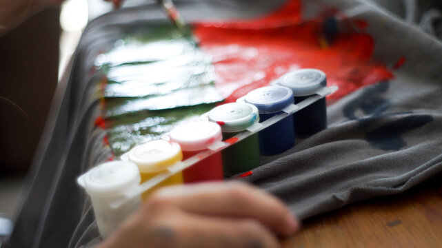 Child hands painting with acrylic colors on a grey t-shirt at home workspace