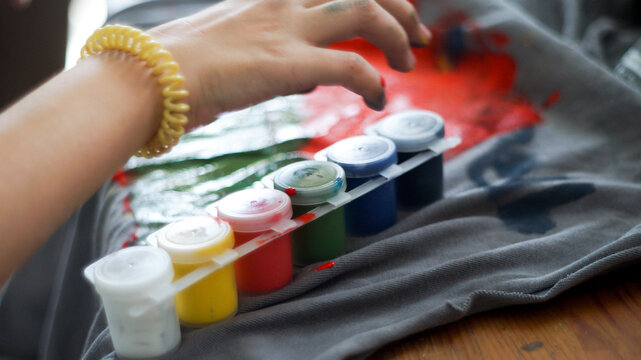 Child hands painting with acrylic colors on a grey t-shirt at home workspace