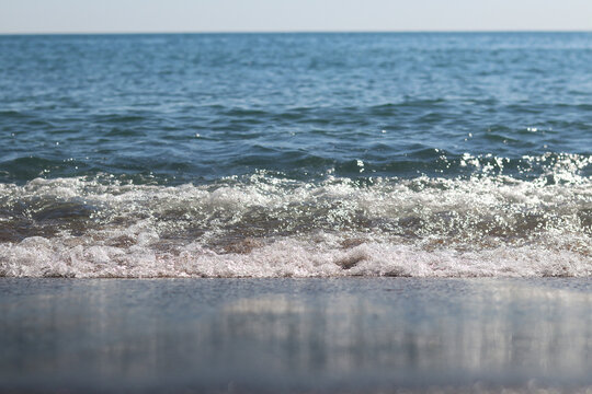 Close-up of a transparent sea wave crashing onto a sandy beach. The sun sparkles on the water surface. Sea surf on a sunny day. Transparent clean water of the Black Sea. Atmosphere of summer vacation