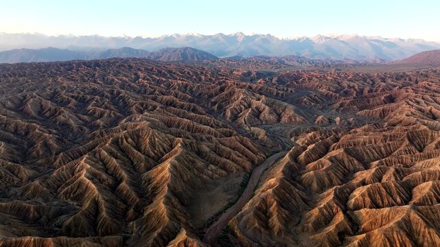 Aerial view of rugged eroded canyons and desert landscape with snow-capped mountains in the distance under golden hour light in Ak Say, Issyk-Kul Region, Kyrgyzstan.