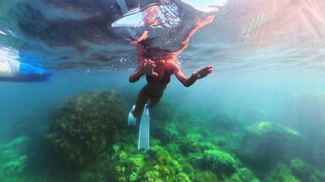 Freediving adventure, a swimmer wearing fins glides past a vibrant green reef under bright sunbeams. Tracking shot.