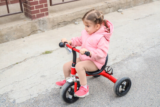 Young Child Rides Red Tricycle Along Rough Sidewalk Beside Brick Building, Wearing Pink Jacket, Beige Leggings, And Sneakers, Showing Early Cycling Practice, Balance, Coordination, Outdoor Play