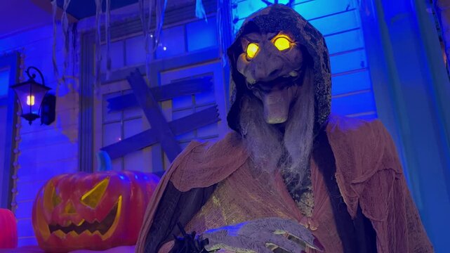 A spooky hooded witch with glowing yellow eyes stands beside a carved pumpkin in front of a haunted house for Halloween.