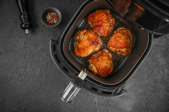 Air fryer with crispy golden-brown chicken thighs inside, accompanied by a small bowl of mixed peppercorns on a textured black surface