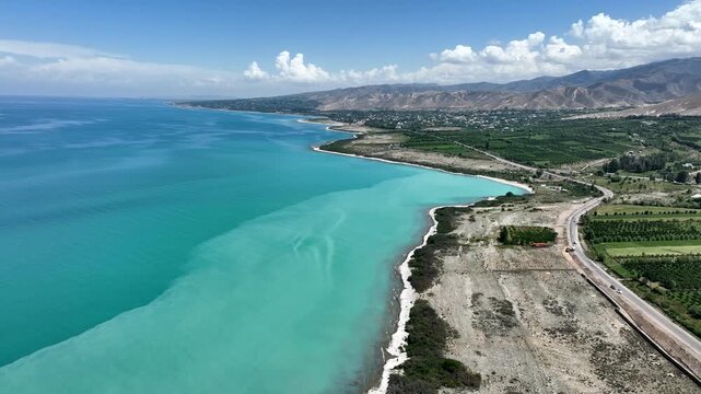Aerial view of the turquoise waters of Issyk Kul lake, with a coastal road and green fields along the shoreline in Cholpon Ata, Issyk-Kul Region, Kyrgyzstan.
