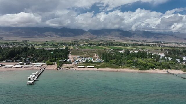 Aerial view of the Issyk Kul lake shoreline with a wooden pier extending into turquoise water, surrounded by green trees and distant mountains in Cholpon Ata, Issyk-Kul Region, Kyrgyzstan.