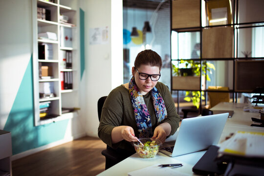 Woman eating salad while working on laptop in modern office