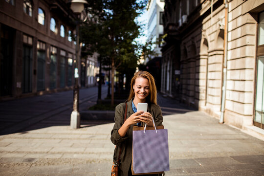 Smiling young woman using smartphone with shopping bag on city street