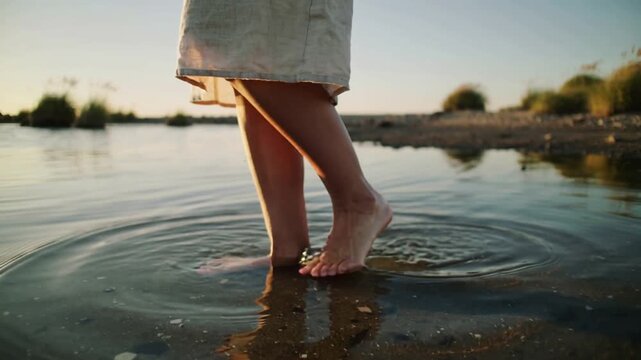 Walking person's lower legs and feet touching shallow shore water, making ripples, dress hem moving