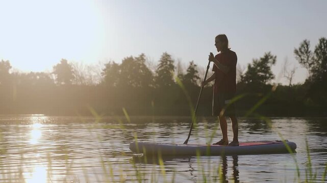 Sup board boad man. A man paddles on a calm lake, the man enjoying the gentle waves beneath his paddle. The setting sun casts a warm glow over the peaceful lake scene.