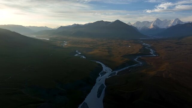 Aerial view of Sary Jaz river winding through the vast mountain valley at sunset with snow-capped peaks in the distance in Karakol, Issyk-Kul Region, Kyrgyzstan.