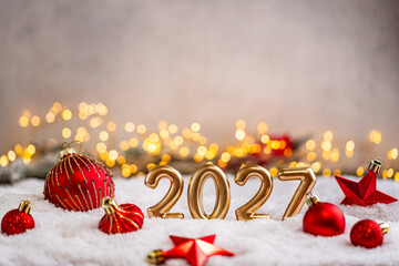 Naklejka premium A Christmas background featuring gleaming gold 2027 balloon numbers surrounded by red baubles, ribbon, and a star on snow, softly lit by warm twinkling lights with copy space.