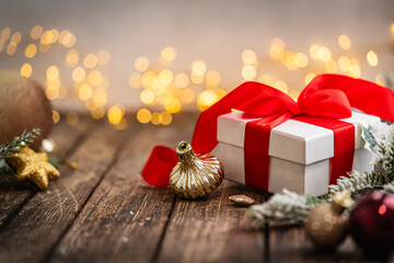 Naklejka premium A Christmas background featuring a classic white gift box tied with a bright red satin bow beside a golden ornament and frosted pine on rustic wood, warmly lit with twinkling lights.