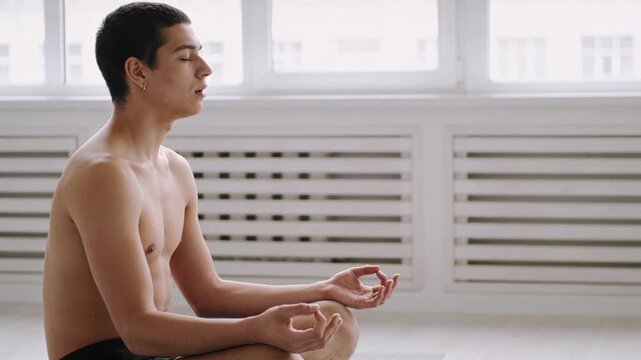 Sunlight drawing shirtless man in shorts wearing hoops breathing slowly beside windows, copy space
