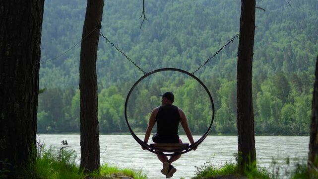 Inspiring static footage from a back view of a male traveler in a black tank top swinging slowly on a circular hanging swing over the Katun River in the Altai Mountains, Siberia.